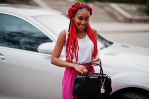 mulher de negócios americano africano na moda em calças cor de rosa e dreads vermelhos posou ao ar livre com bolsa contra seu carro de luxo. foto