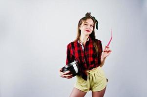 jovem dona de casa engraçada na camisa quadriculada e short amarelo pin up estilo com panela e colher de cozinha isolado no fundo branco. foto