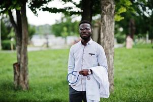 jovem médico americano africano segura jaleco branco por lado com um estetoscópio posado ao ar livre. foto