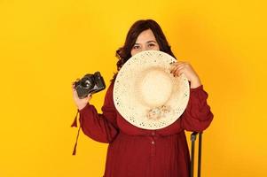 mulher atraente viajante do sul da Ásia no vestido vermelho escuro, chapéu posou no estúdio em fundo amarelo com mala amd velha câmera fotográfica vintage. foto