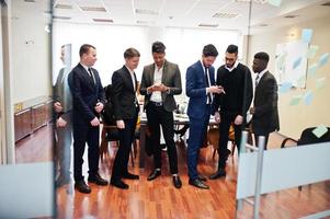 seis homens de negócios multirraciais em pé no escritório e usam telefones celulares. grupo diversificado de funcionários do sexo masculino em trajes formais com telefones celulares. foto