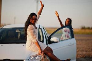 duas mulheres negras muito jovens usam pose de vestido de verão no carro contra um campo de girassol. foto