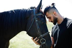 homem árabe de barba alta usa preto com cavalo árabe. foto