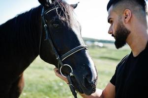 homem árabe de barba alta usa preto com cavalo árabe. foto