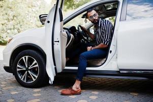 homem árabe bem sucedido usa camisa listrada e óculos de sol posam ao volante de seu carro suv branco. homens árabes elegantes no transporte. foto