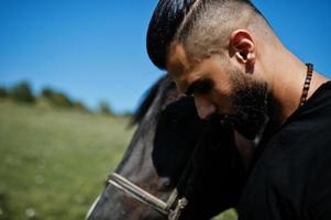 homem árabe de barba alta usa preto com cavalo árabe. foto