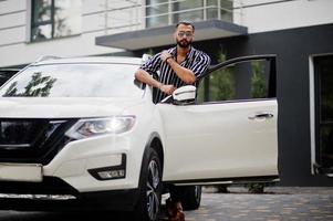 homem árabe bem sucedido usa camisa listrada e óculos de sol posam perto de seu carro suv branco. homens árabes elegantes no transporte. foto