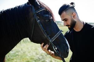 homem árabe de barba alta usa preto com cavalo árabe. foto