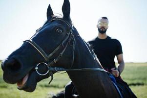 homem de barba alta árabe usa preto e óculos de sol montam cavalo árabe. foto