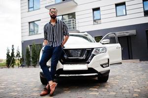 homem árabe bem sucedido usa camisa listrada e óculos de sol posam perto de seu carro suv branco. homens árabes elegantes no transporte. foto