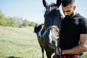 homem árabe de barba alta usa preto com cavalo árabe. foto