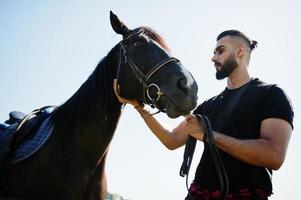 homem árabe de barba alta usa preto com cavalo árabe. foto