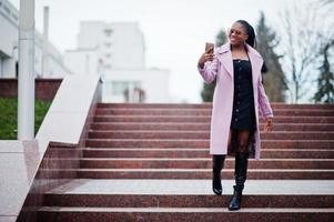 jovem elegante linda afro-americana na rua contra escadas, vestindo casaco de roupa de moda, óculos com telefone celular nas mãos. foto