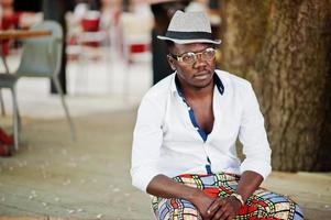 homem afro-americano elegante na camisa branca e calça colorida com chapéu e óculos posou ao ar livre. menino modelo preto elegante. foto