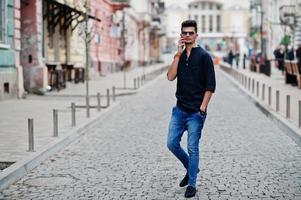 homem elegante modelo indiano em roupas casuais, camisa preta e óculos de sol posou ao ar livre na rua da índia e falando ao telefone. foto