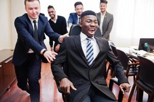 seis homens de negócios multirraciais em pé no escritório e rolar o homem na cadeira. grupo diversificado de funcionários do sexo masculino com roupa formal se divertindo. foto
