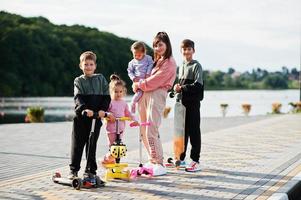 jovem mãe elegante com quatro filhos ao ar livre. família de esportes passa o tempo livre ao ar livre com patinetes e patins. foto