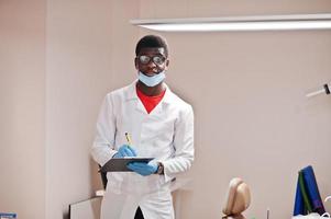 médico americano africano em óculos e máscara posou na clínica com prancheta preta à mão e escrevendo algo. foto