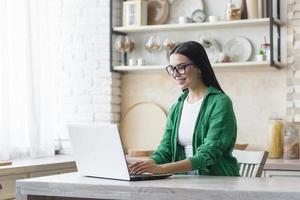 jovem mulher de negócios linda de óculos e camisa verde trabalhando em casa na cozinha foto