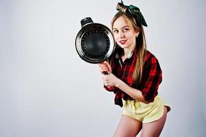 jovem dona de casa engraçada na camisa quadriculada e short amarelo pin up estilo com frigideira isolada no fundo branco. foto