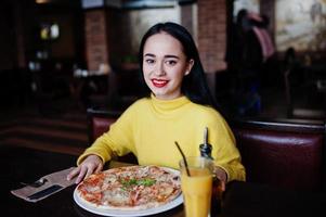 menina morena engraçada de suéter amarelo comendo pizza no restaurante. foto