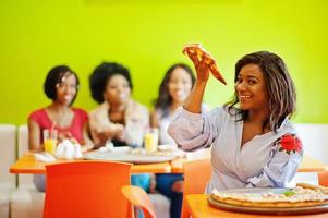 mulher africana com pizza sentado no restaurante contra meninas de pele escura. foto