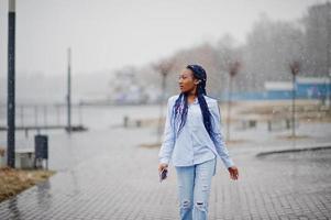 menina afro-americana elegante com dreads segurando o celular na mão, ao ar livre com tempo de neve. foto