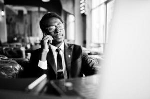 homem afro-americano de negócios usa terno preto e óculos sentado no escritório e falando no telefone atrás do laptop. foto