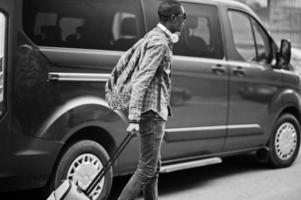 homem afro-americano na camisa quadriculada, óculos escuros e fones de ouvido com mala e mochila. viajante negro contra carro de van. foto