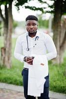 jovem médico americano africano segura jaleco branco por lado com um estetoscópio posado ao ar livre. foto