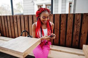 garota afro-americana na moda em calças cor de rosa e dreads vermelhos posou ao ar livre com telefone celular. foto