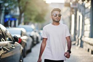 homem de barba indiana elegante em óculos de sol e camiseta rosa. modelo da índia andando ao ar livre por do sol nas ruas da cidade. foto