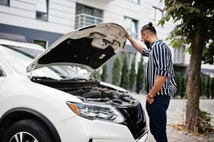 homem árabe bem sucedido usa camisa listrada e óculos de sol posam perto de seu carro suv branco, verifique o motor com o capô aberto. foto