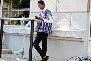 homem africano em roupas tradicionais e óculos no caffe ao ar livre, bebe café e procura notícias diárias no telefone. foto