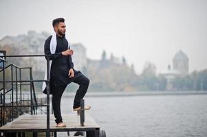 homem elegante indiano em roupas tradicionais pretas com lenço branco posou ao ar livre contra cais nebuloso do mar. foto