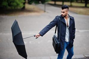 homem de barba árabe alto na moda usa casaco preto com guarda-chuva e bolsa posada no dia de chuva. foto