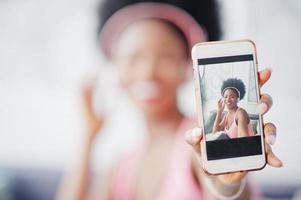 foto da tela do telefone móvel jovem afro-americana sentada no sofá, ouvir música em fones de ouvido.