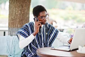homem africano em roupas tradicionais e óculos sentado atrás do laptop no caffe ao ar livre e falando no celular. foto