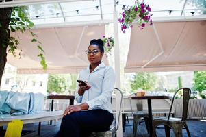 garota afro-americana usa óculos com celular sentado no caffe ao ar livre. foto