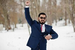 homem de negócios de barba indiana elegante de terno e óculos de sol posou em dia de inverno ao ar livre, mãos para cima e emoções felizes. foto