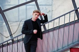 jovem macho macho elegante na jaqueta preta posou ao ar livre da rua. homem modelo incrível no tonel de escadas vermelhas. foto