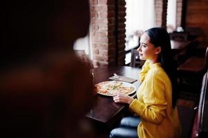 menina morena engraçada de suéter amarelo comendo pizza no restaurante. foto