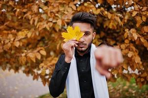 homem elegante indiano em roupas tradicionais pretas com lenço branco posou ao ar livre contra o outono amarelo e mostra o dedo. foto