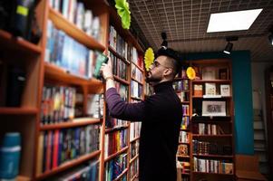 homem alto e inteligente estudante árabe, use gola rulê violeta e óculos, na biblioteca selecionando livro nas prateleiras.