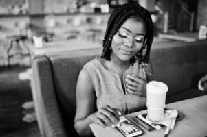 mulher afro-americana atraente sentada à mesa no café com café com leite e olhando no celular. foto