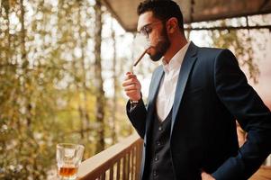 bonito homem árabe bem vestido fuma charuto na varanda do pub. foto