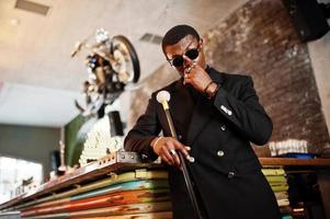 elegante cavalheiro afro-americano na elegante jaqueta preta e óculos de sol, segurando a bengala retrô como balão de cana ou bengala com alça de bola de diamante dourado. homem afro elegante rico. foto