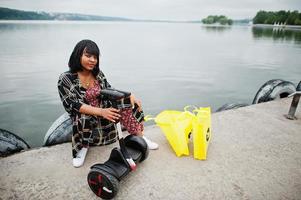 linda mulher afro-americana sente-se perto de segway ou hoverboard. garota negra com símbolo de reciclagem de sacos ecológicos de pano amarelo. foto