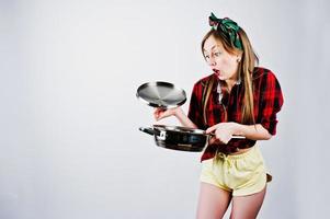 jovem dona de casa engraçada na camisa quadriculada e short amarelo pin up estilo com frigideira isolada no fundo branco. foto