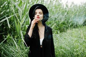 garota fumante sensual toda em chapéu e lábios pretos, vermelhos. mulher dramática gótica fumando na cana comum. foto